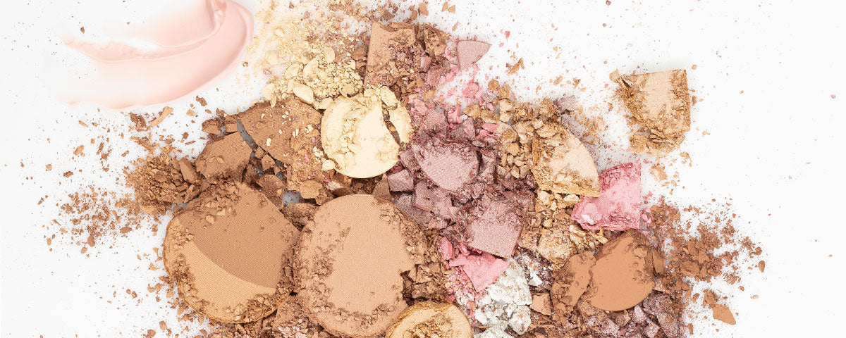 Crushed powder makeup pans in browns, pinks and golds with a smear of pink cream product on a white background.