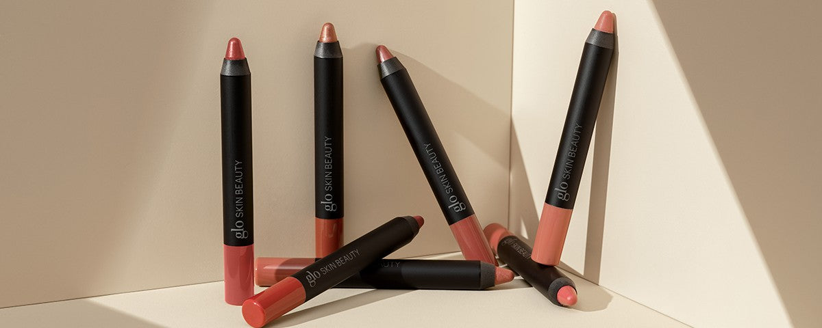 Five nude lip crayons on a beige backdrop, black barrels labeled 'glo SKIN BEAUTY'.