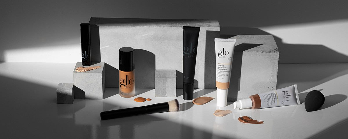 Front studio shot of Glo Skin Beauty foundations and moisture tints with shade swatches, brush, and sponge on concrete blocks