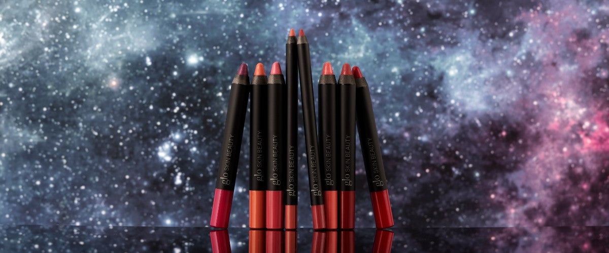 Nine red and pink lip crayons labeled "glo SKIN BEAUTY" standing on a reflective surface with a starry galaxy background