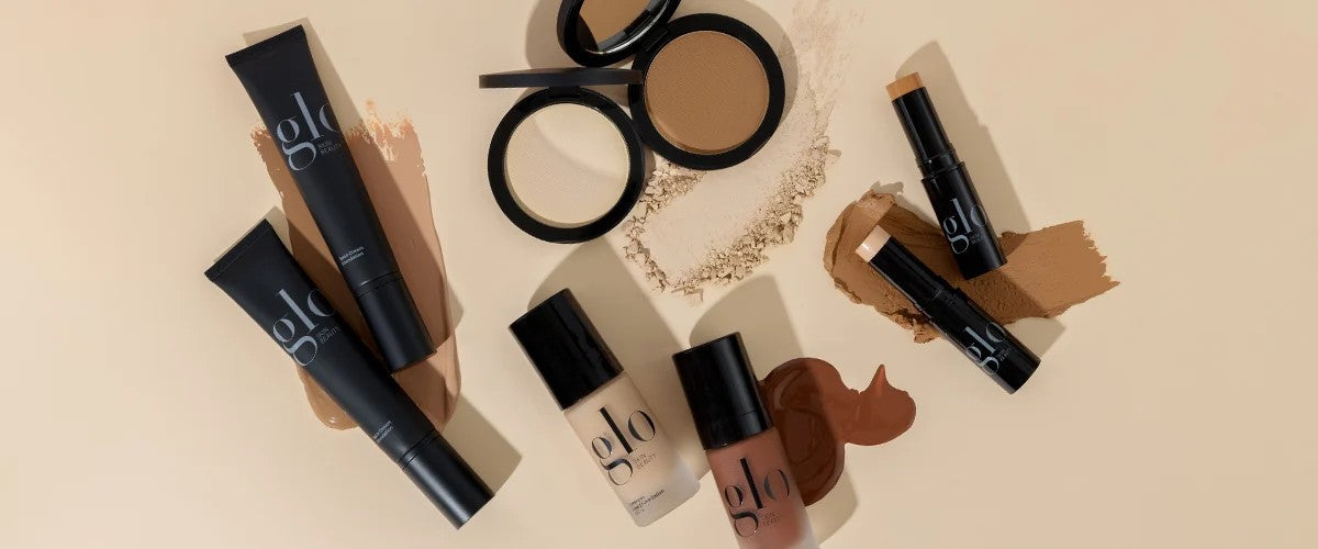 Assortment of glo foundation products - tubes, liquid and stick foundations, compacts and powder swatches on a beige background