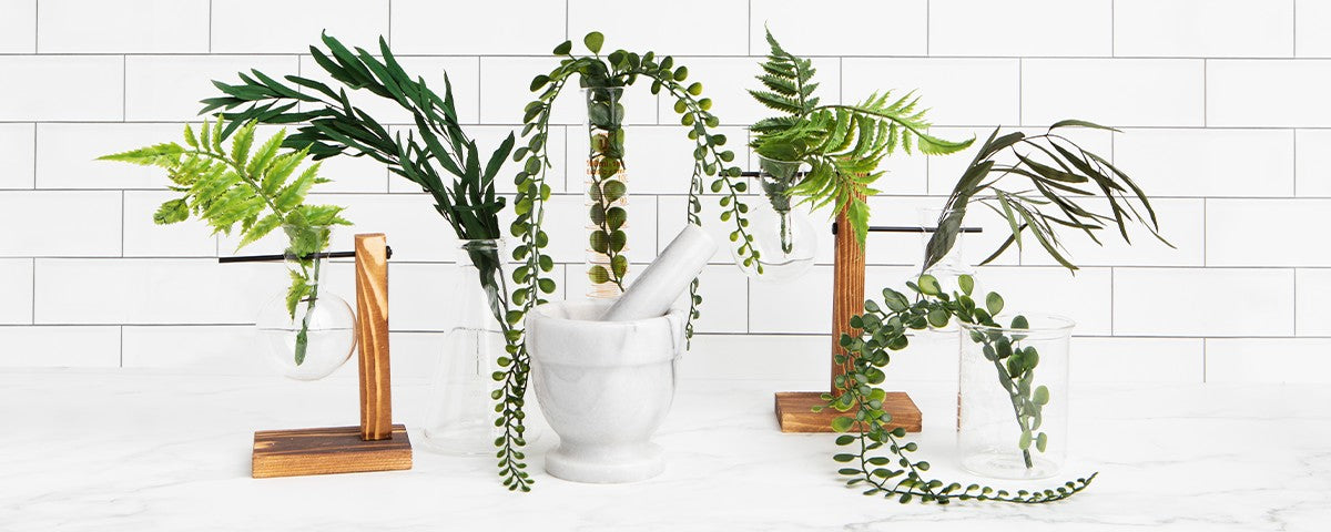 Marble mortar and pestle with ferns in glass vases and hanging test tubes on a white tiled countertop.