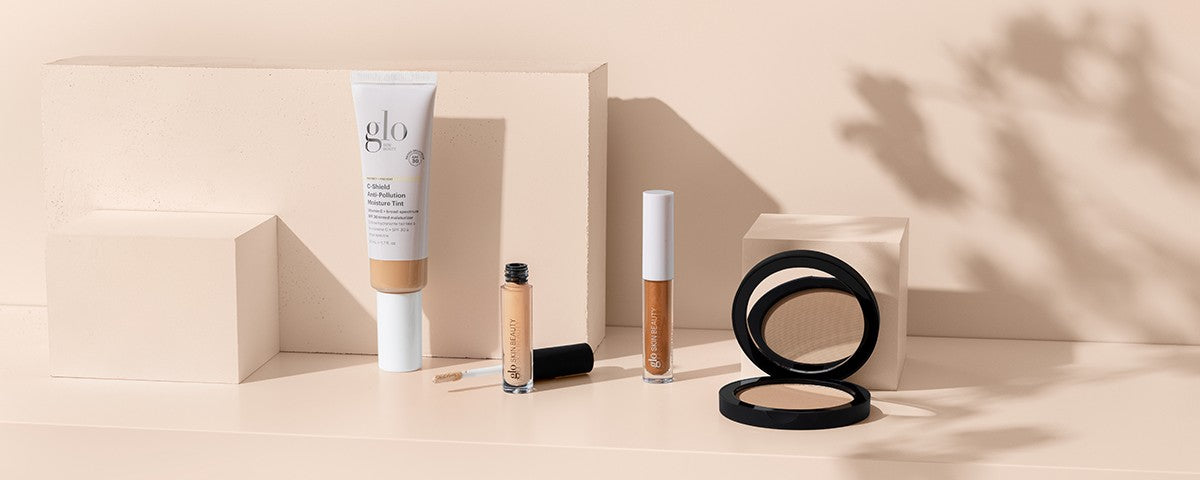 glo Skin Beauty products - moisture tint tube, concealer, lip gloss, and compact powder arranged on beige blocks