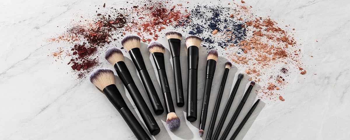 Black-handled makeup brushes fanned on marble with scattered powdered eyeshadow and blush pigments