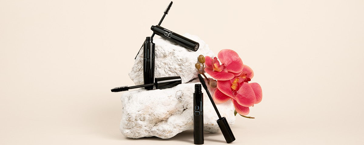Three black mascara tubes with wands on white stones beside a pink orchid
