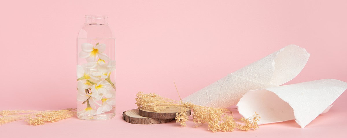 glass bottle of water with white plumeria flowers beside wood slices and dried grasses on pink background