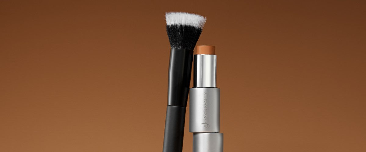 Dense angled makeup brush beside a stick foundation on a warm brown gradient background.