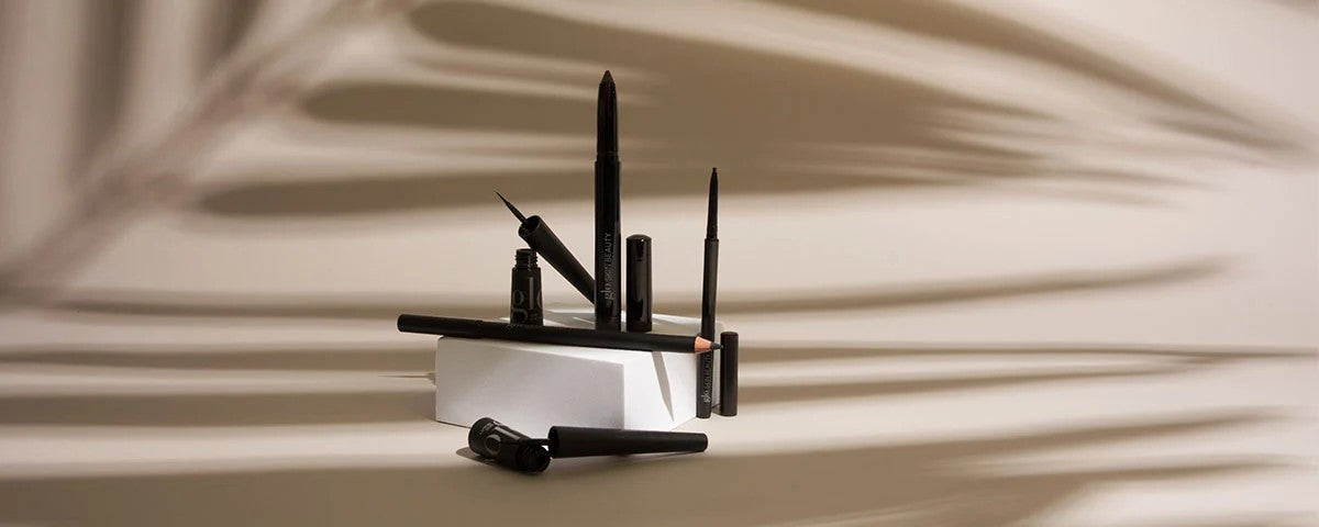Assorted black eyeliners and a sharpened pencil balanced on a white block with palm-leaf shadows on a beige background