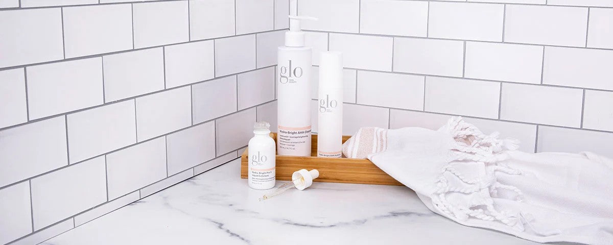Three white glo skincare bottles on a wooden tray beside a white towel on a tiled bathroom counter.