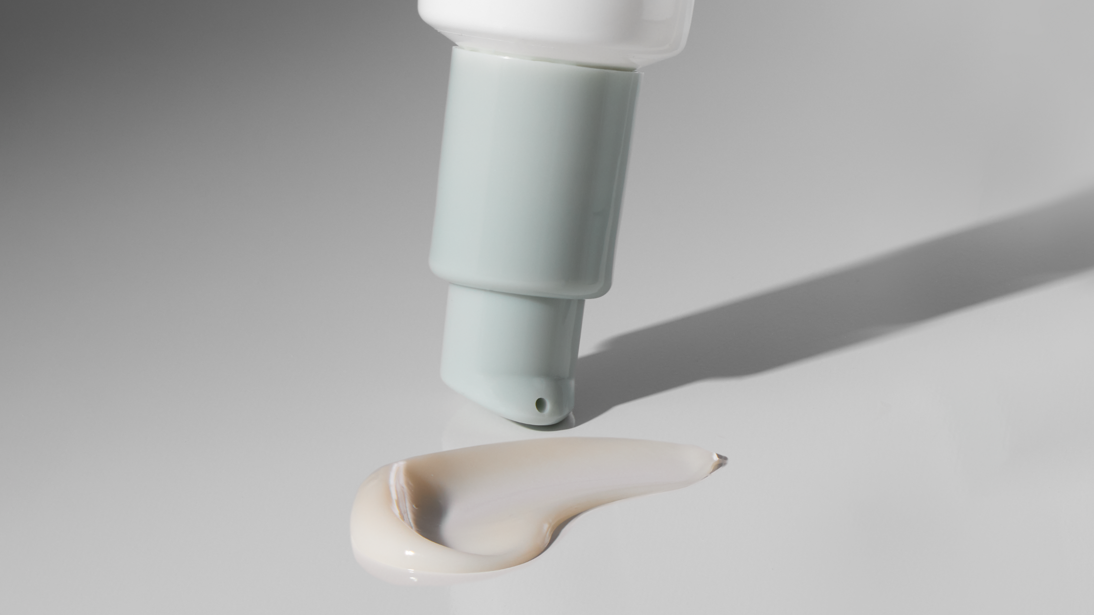 Cream dispenser pump with pale green nozzle tilted above a beige skincare cream swatch on a glossy surface