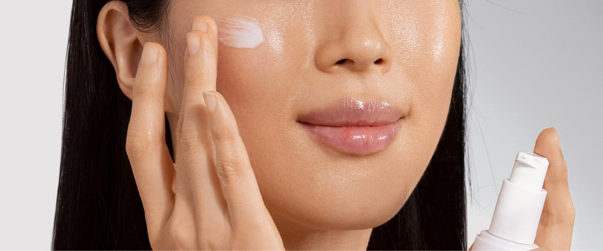 Woman applying moisturizer to her cheek with fingers; white pump bottle visible at right.