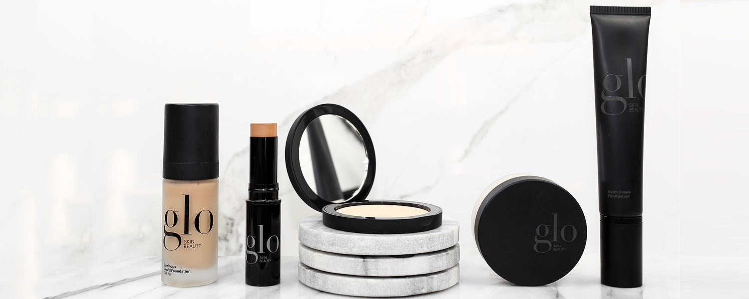 Five glo SKIN BEAUTY products on marble: Luminous Liquid Foundation, Satin Cream Foundation, stick, compact with mirror