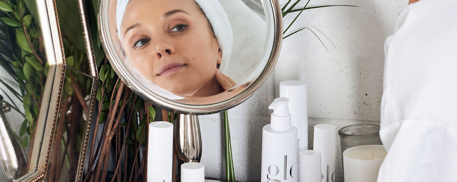 Woman with towel on head checking her cheek in a round vanity mirror, surrounded by white 'glo' skincare bottles and a candle.