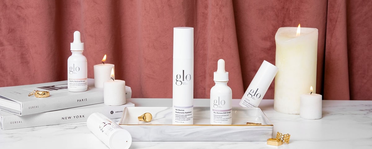 Glo skincare products and dropper bottles on a marble tray with candles and stacked books labeled CEREAL, PARIS, NEW YORK