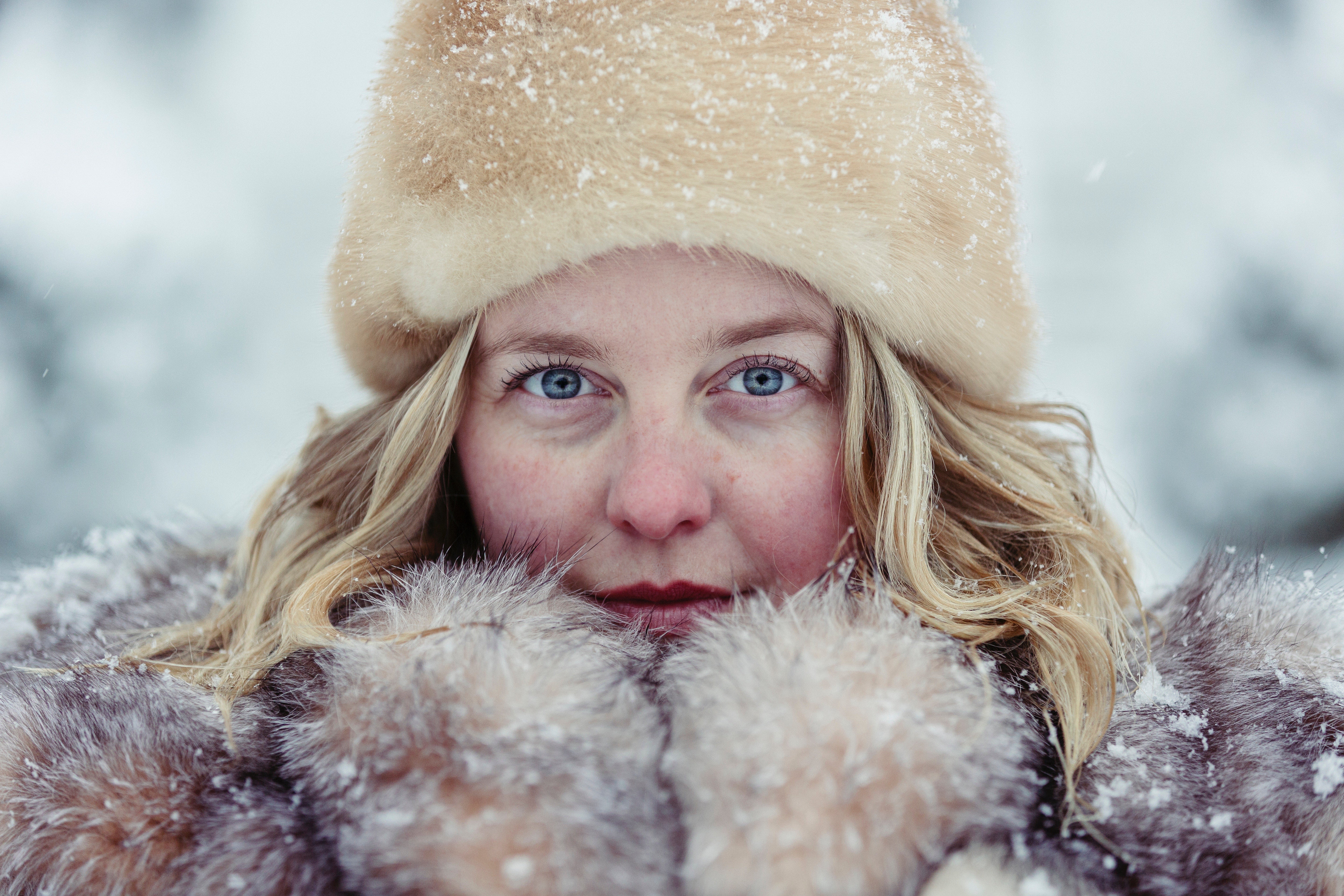 Close-up of woman's face with blue eyes, rosy cheeks, fur collar and beige hat dusted with snow