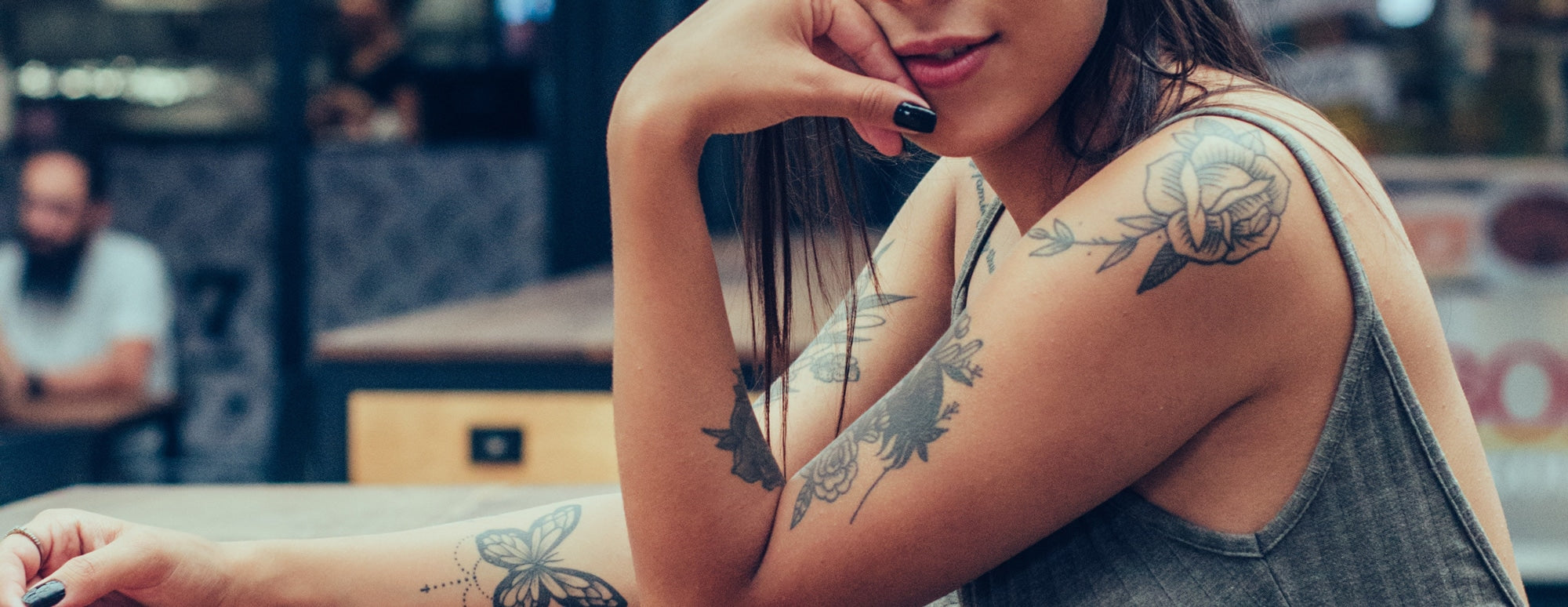 Young woman seated at a table, forearm butterfly and floral tattoos and a rose tattoo on her upper arm