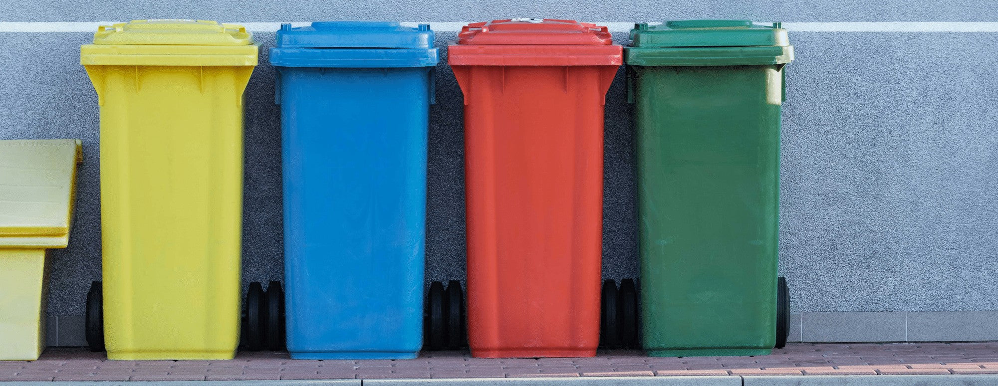 Four outdoor wheeled trash bins in yellow, blue, red, and green lined up against a gray wall.