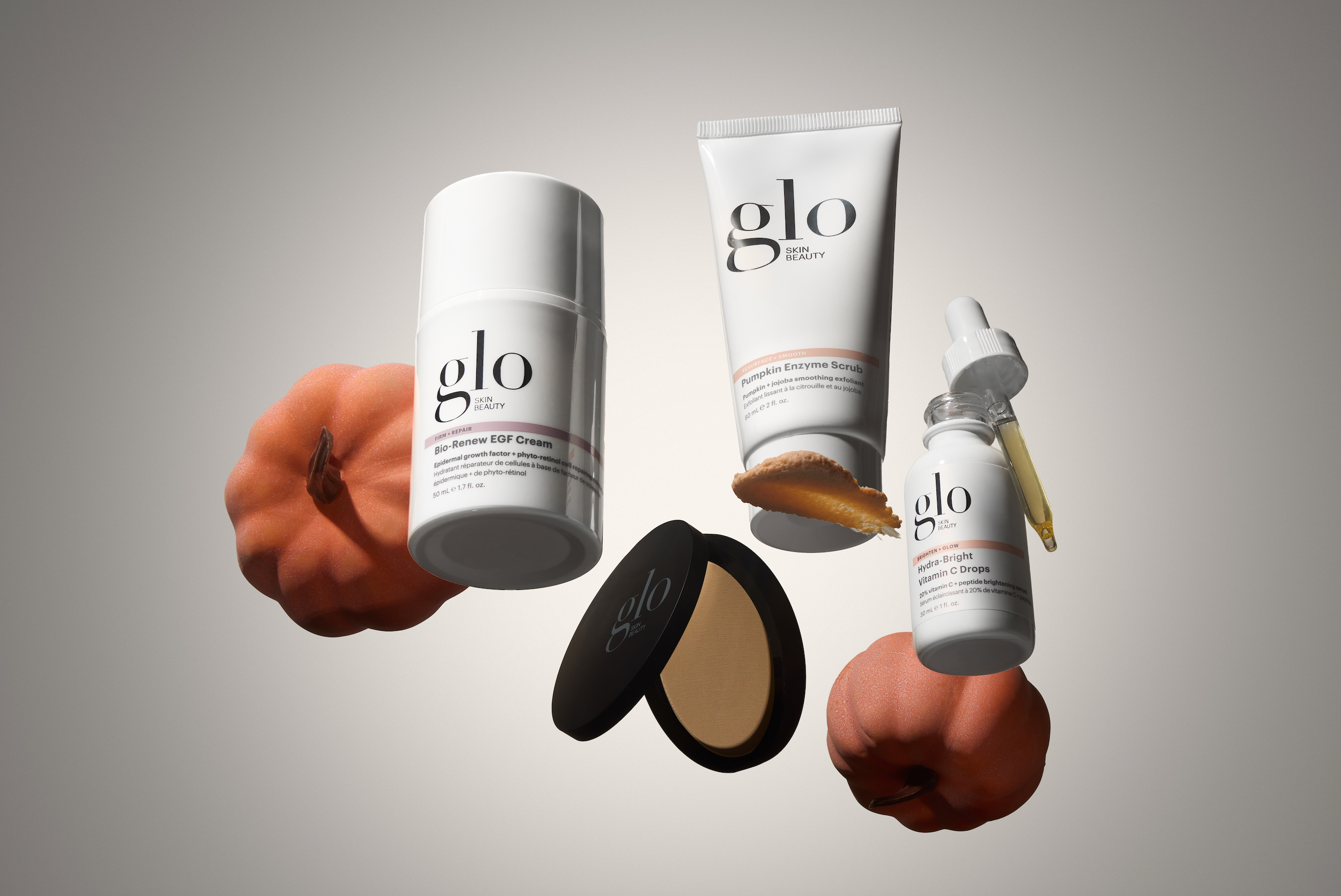 Angled view of Glo Skin products floating: EGF cream, pumpkin scrub smear, compact powder, and C drops on neutral background