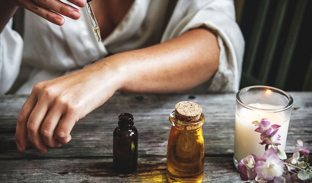 Person using a dropper to apply oil to their forearm, two small bottles and a candle on a wooden table