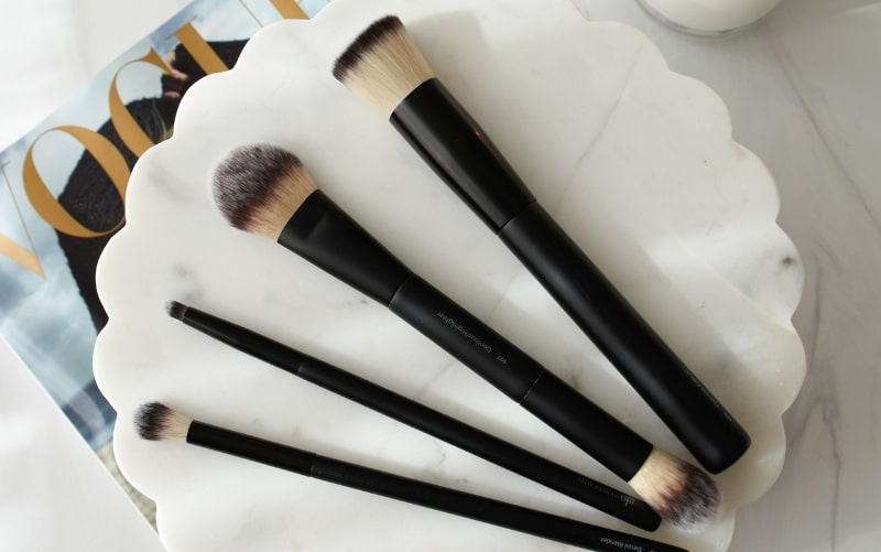 Five black-handled makeup brushes fanned on a scalloped white marble tray over a VOGUE cover