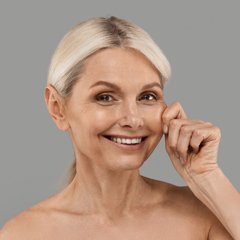 Smiling mature woman pinching her cheek to show skin firmness against a neutral background