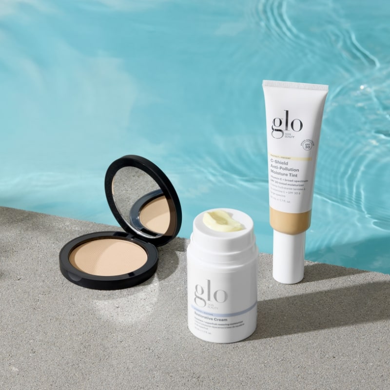 Three glo Skin Beauty products — compact powder, cream stick, and moisture tint tube on poolside concrete with blue water background