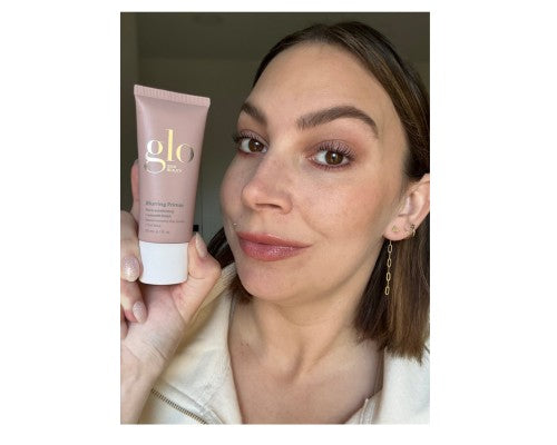 Woman holding a tube labeled glo Blurring Primer next to her face to show finished makeup and skin texture.