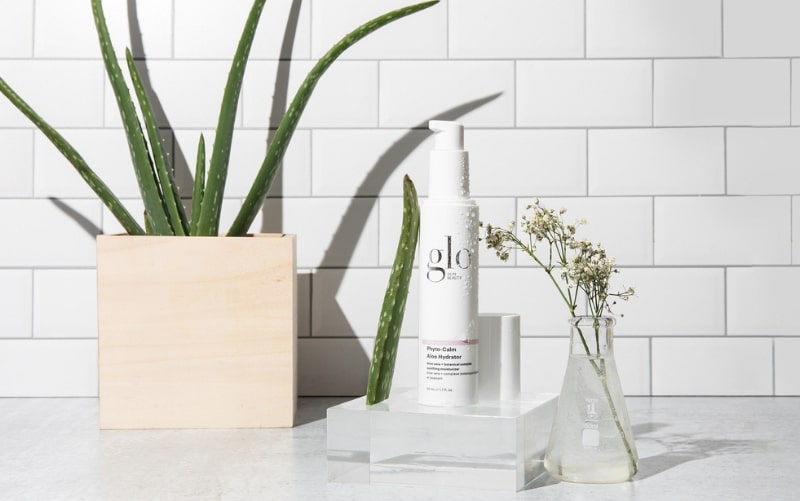 glo bottle 'Phyto-Calm Aloe Hydrator' with aloe leaf, potted aloe and glass flask of flowers on subway-tile background.