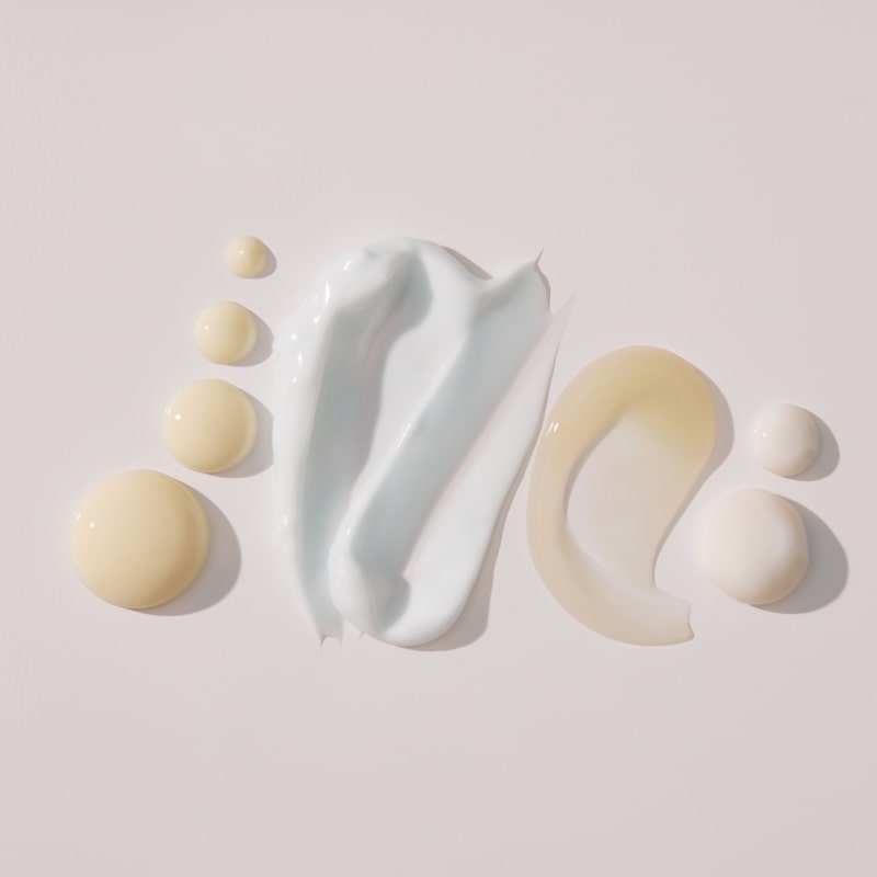 Skincare swatches and droplets: pale blue and white creams, beige lotion, and translucent gel on a pale background.