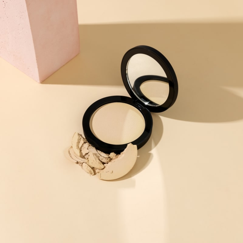 Compact pressed powder in a black case with mirror, crushed powder pieces beside it on a beige surface