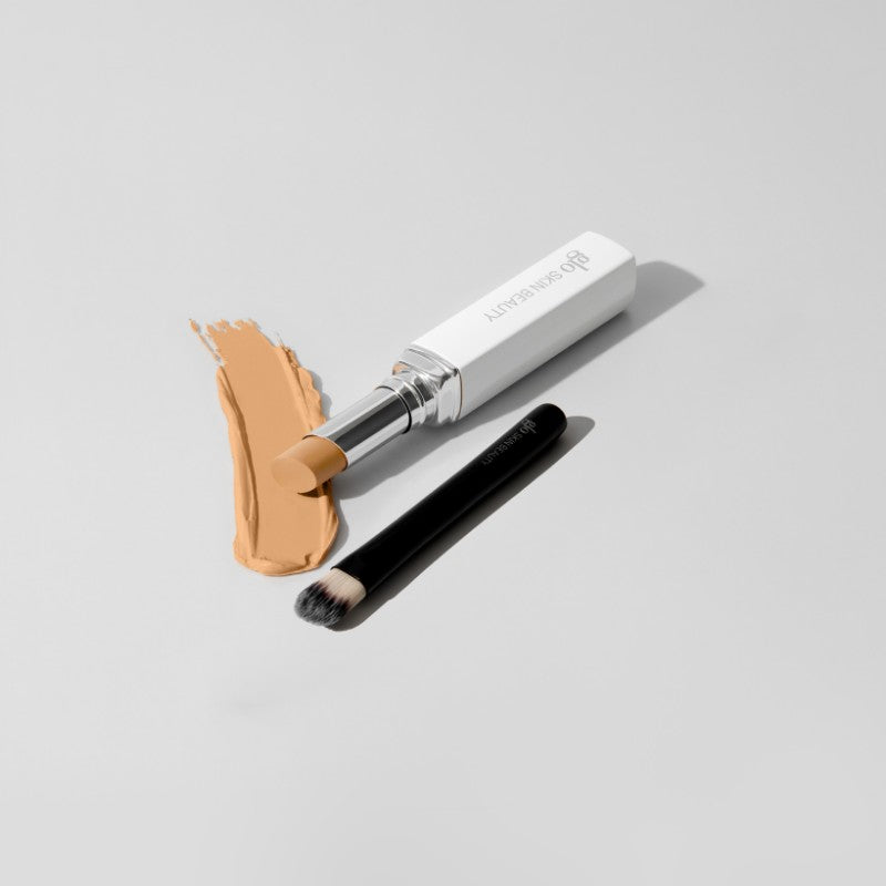 Stick concealer labeled 'DB SKIN BEAUTY' beside a small blending brush and a swatch of medium beige concealer.