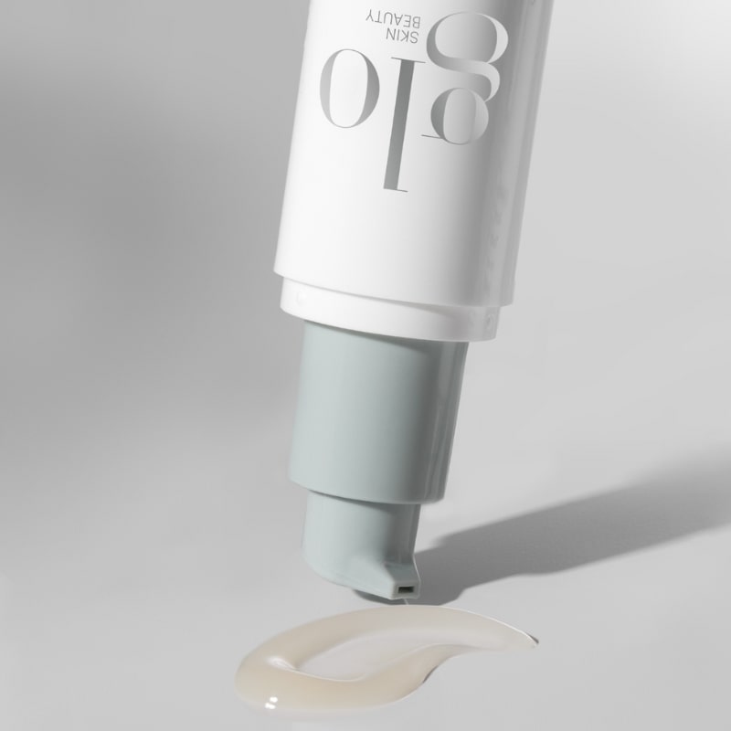 Upside-down glo SKIN BEAUTY pump tube dispensing a pale cream drop onto white surface.