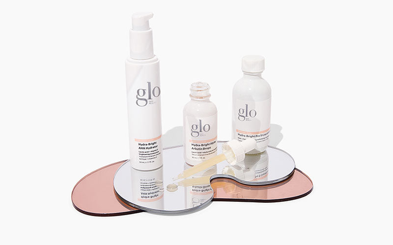 Three glo Hydra-Bright AHA skincare bottles, one with a dropper, on mirrored pink trays