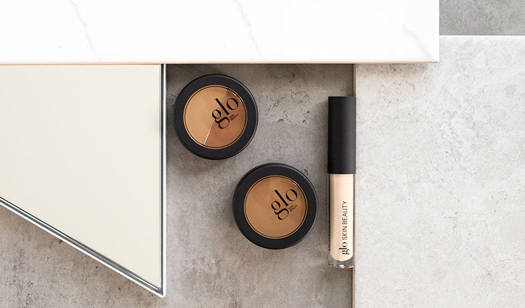 Two brown concealer compacts with 'glo' logo and a concealer tube labeled 'glo SKIN BEAUTY' on a stone surface.