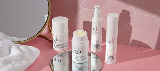Three white glo skincare bottles and an open jar of cream on a round mirror, pink hexagon-tile backdrop.
