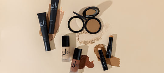 Assorted glo-branded makeup: tubes, foundation bottle, compact and stick with swatches on beige background.