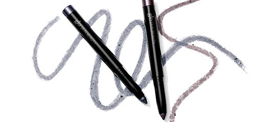 Two black eyeliner pencils with gray and mauve swatches sketched behind them