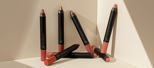 Six glo skin beauty lip crayons in pink and nude shades arranged on a beige corner surface.