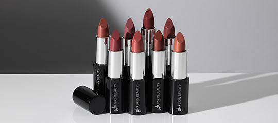 Assorted mauve and red lipsticks in black tubes labeled glo SKIN BEAUTY displayed on a reflective gray surface