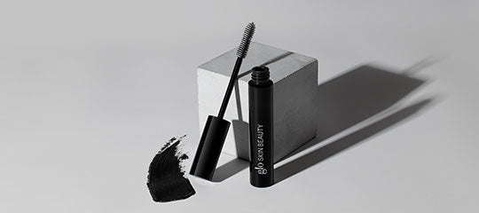 Black mascara tube labeled 'glo SKIN BEAUTY' with wand, a smear of mascara, and a long shadow on a gray background.