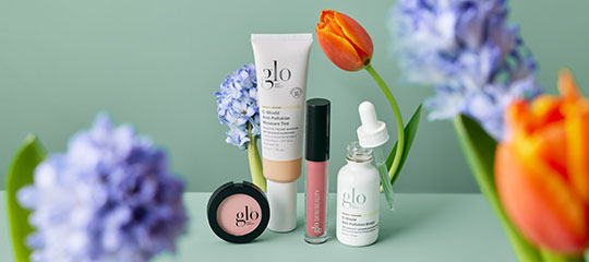 glo skincare products — compact, tinted tube, lip gloss and dropper serum staged with tulips and hyacinths on green background.