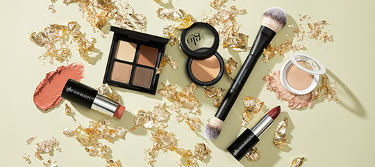 Assortment of makeup: eyeshadow quad, pressed powders, brush, two lipsticks and scattered gold leaf on pale background