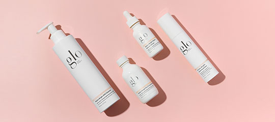 Four white bottles labeled glo (skincare packaging) arranged diagonally on a pink background with cast shadows.