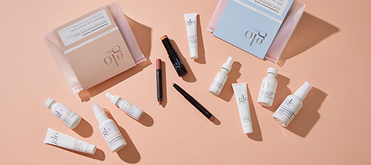 Assorted glo skincare bottles, tubes and makeup pencils arranged on a peach background.