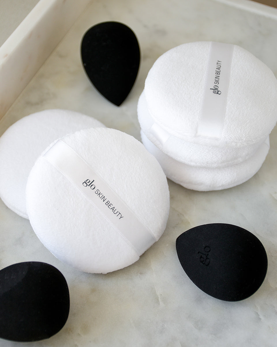 Round white makeup puffs with 'glo SKIN BEAUTY' ribbon and black teardrop sponges embossed 'glo' on a marble tray.