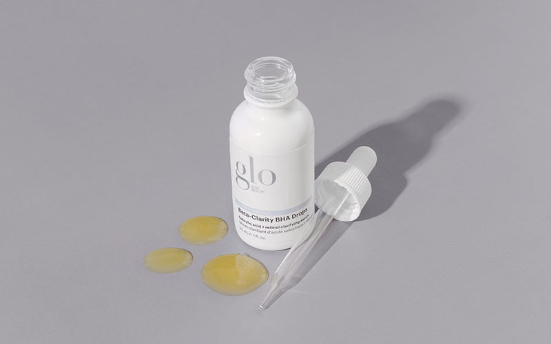 glo bottle labeled 'Beta-Clarity BHA Drops' with pipette and three yellow serum drops on gray background