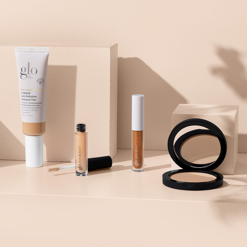Beige flatlay of glo SKIN BEAUTY products: C-Shield Anti-Pollution Moisture Tint SPF 30, concealer, lip gloss, compact.