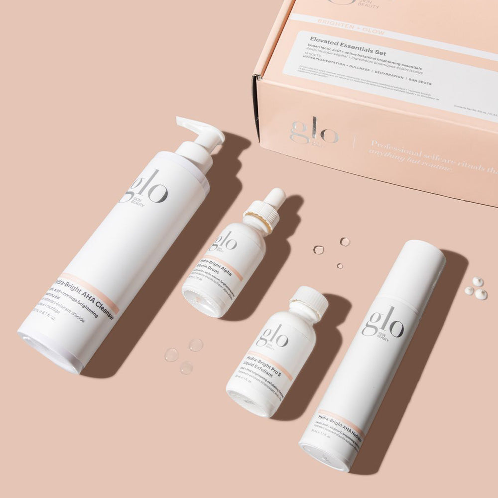 glo skincare set with four white bottles and a box on peach background; reads Brighten + Glow and Elevated Essentials Set