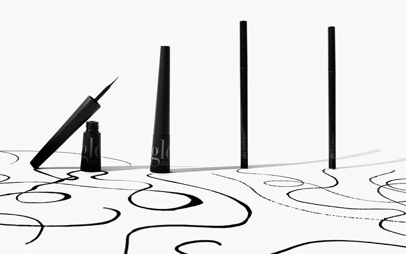 Black liquid eyeliner bottle with applicator and three upright 'glo' pens on white background with black ink swirls