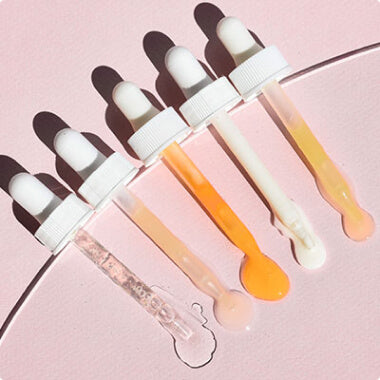 Five skincare droppers with varying serum colors—clear, pale pink, peach, orange, and cream—on a pale pink background.